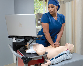 A female nurse in blue scrubs and cap performs CPR compressions on an adult RQI manikin. An infant manikin rests on a table next to a laptop displaying training material.