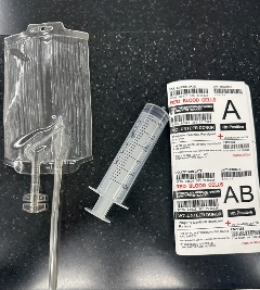 Clear empty blood bag, a large clear syringe, and two blood-type labels marked A and AB lying on a black speckled surface.