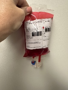 A hand holds a red-filled simulated blood bag against a white wall, with a printed label showing blood type options, barcodes, and tubing attached at the bottom.