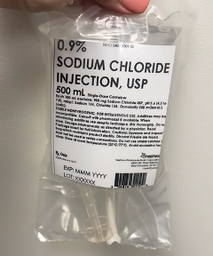 A hand holds a clear 500 mL bag of 0.9% sodium chloride injection with printed labeling, tubing visible inside the package, against a white background.