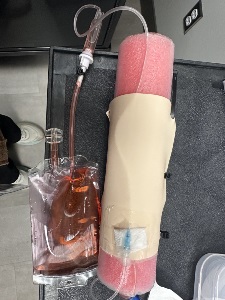 A simulated IV setup with a pink foam arm wrapped in a beige sleeve, an IV catheter taped in place, and tubing connecting to a clear bag filled with red liquid, all arranged on a dark surface.