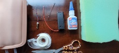 Supplies on a table, including wires, a black battery holder, a bottle of silicone glue, clear tape in a dispenser, scissors with patterned handles, foam, and a intravenous/venipuncture practice pad