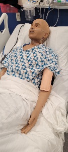 A manikin lies in a hospital bed wearing a patterned gown, with the wearable AV fistula strapped to the arm  and medical equipment visible in the background