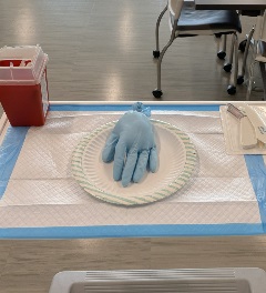 A tabletop setup with a blue glove placed on a paper plate atop absorbent pads. A red sharps container sits to the left, and packaged medical supplies are on the right. Classroom chairs and tables are visible in the background.