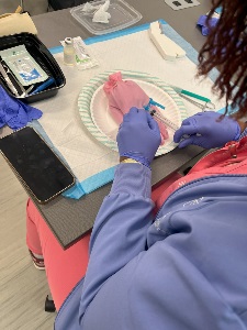 A person wearing purple gloves practices an injection on a pink glove placed on a paper plate over absorbent pads. Medical supplies, a syringe, and an open kit are on the table, with a phone beside them.