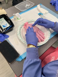 A person wearing purple gloves practices an injection on a pink glove placed on a paper plate over absorbent pads. Nearby are medical supplies, a syringe, a vial, an open kit, and a phone on the table