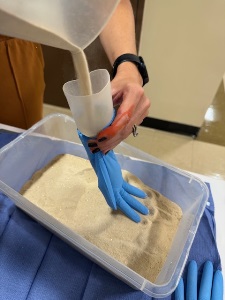 A person pours sand from a clear container into a blue glove positioned in a plastic bin filled with sand, with another blue glove resting nearby on a blue pad.