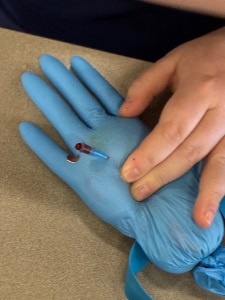 A hand presses on a sand-filled blue glove with a small tube inserted into it, showing a drop of red liquid near the entry point.
