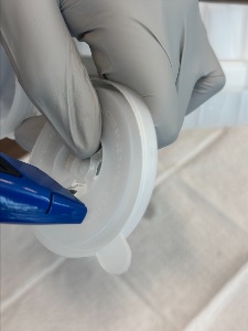 Gloved hands hold a plastic bottle cap while a blue utility knife is used to cut into the center opening, with a medical pad underneath.