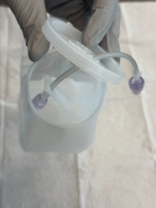 Gloved hands holding an open plastic container lid while IV extension tubing with a purple needleless connector is fed through the port into the container.