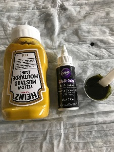 bottle of yellow mustard, bottle of black food coloring, and a medication cup with yellow mustard and black food coloring