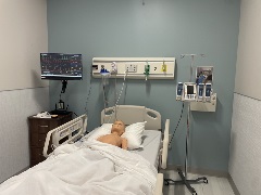 Simulation hospital room with a manikin in a patient bed, bedside monitor displaying vital signs, IV pumps, oxygen and suction equipment, and clinical supplies arranged for a nursing scenario.