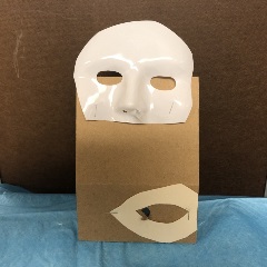 Cardboard with plastic full-face Halloween mask cut below the nose and top of mask stapled to the top of cardboard. Paper cut in teardrop shape with middle cut-out stapled to bottom of cardboard. Cardboard has hole in the middle of the teardrop.