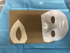 Corrugated cardboard sheet with white plastic full-face halloween mask cut below the nose and top mask stapled to the top of cardboard. White paper cut in shape of a teardrop with middle cut-out stapled to bottom of cardboard.