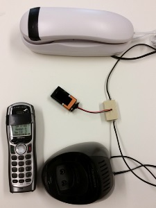 white corded phone wired to the phone coupler with a 9V battery attached by the coupler to a black cordless phone