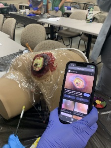 Lifelike wound model on a manikin's buttocks, with a person wearing blue gloves applying wound makeup. A smartphone displays a reference image of a pressure ulcer for accuracy in recreating the wound.