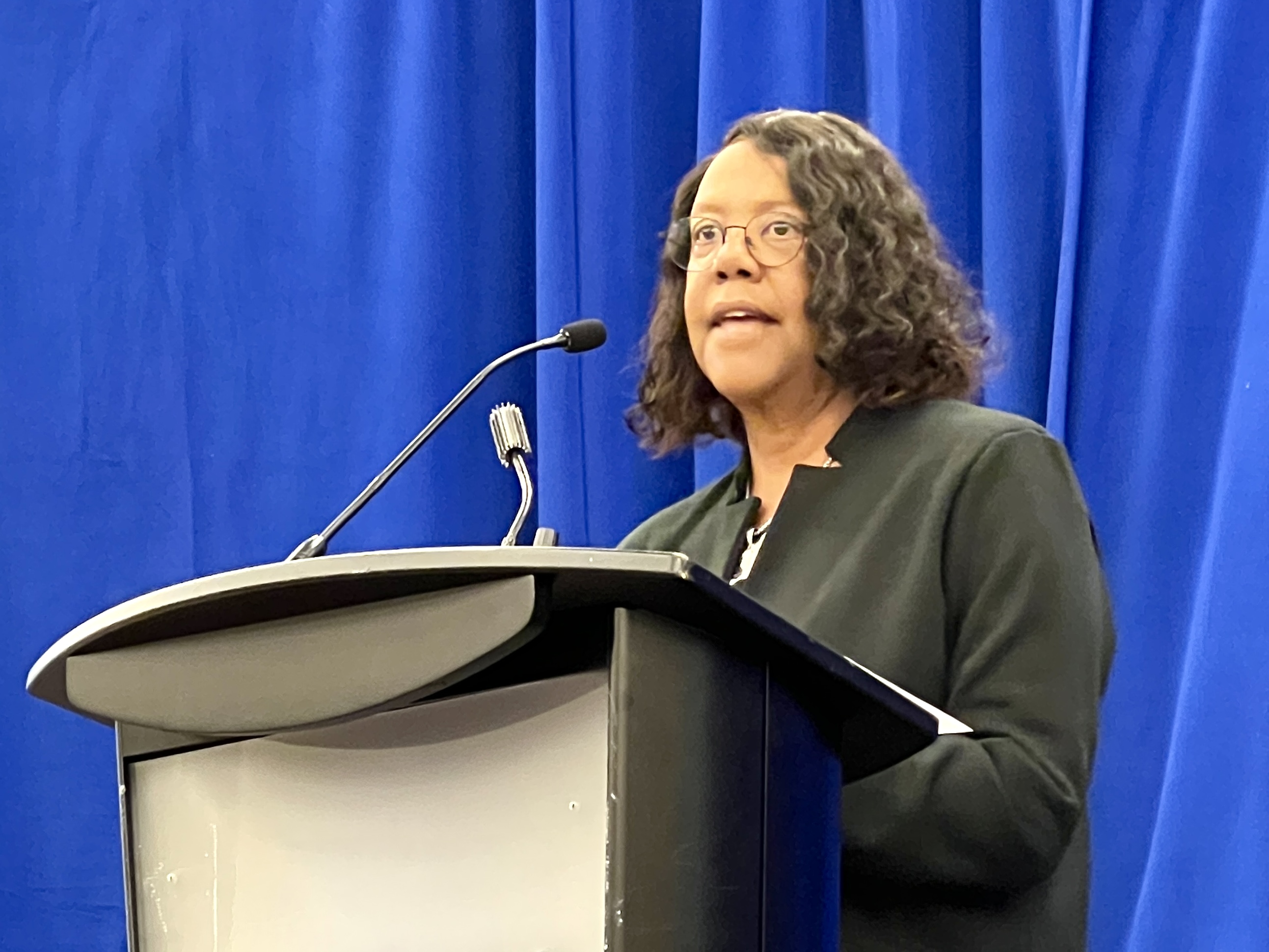 Dr Linda Scott A nurse educator with short dark hair in a black suit speaks from a conference podium