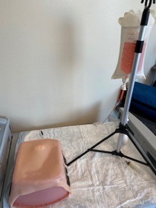 hung IV bag with blood-colored liquid and upside down hospital basin covered with simulated skin on a draped cart