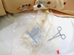 empty 500ml IV bag, empty gallon-size plastic storage bag and hemostat on dressed table next to manikin torso