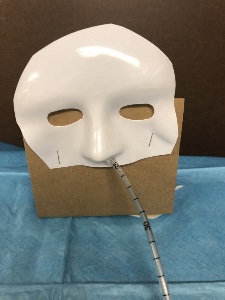 Corrugated cardboard sheet with white plastic full-face halloween mask cut below the nose and top mask stapled to the top of cardboard. Right nostril has nasal tubing running through it.